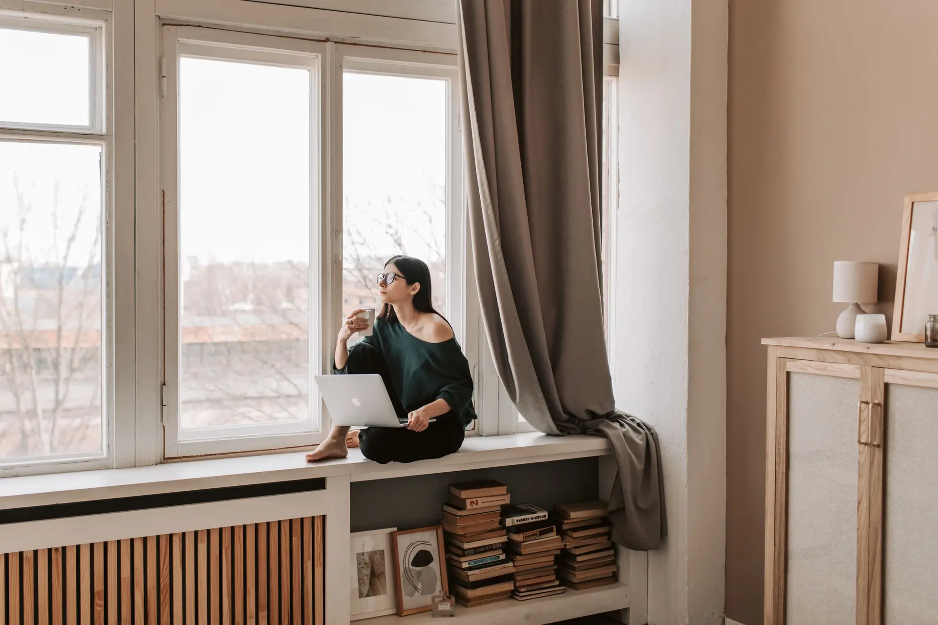 Eine Frau sitzt entspannt mit Tee und Laptop an einem hellen Fenster